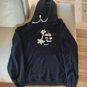 Svart hoodie Axel Arigato - Svart hoodie från Axel Arigato i 100% ekologisk bomull. Stor broderad bokstav och blommor framtill, vita snören i huvan och klassisk känguruficka. Snygg och relaxed passform, perfekt för dig som gillar streetstyle med en twist.