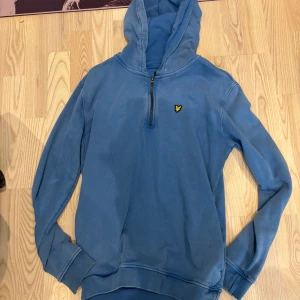 Blå hoodie från Lyle & Scott - Blå hoodie från Lyle & Scott med klassisk gul logga på bröstet. Tröjan har huva, lång ärm och half zip-dragkedja framtill. Perfekt för en chill och avslappnad stil. Tillverkad i mjuk bomull för extra komfort.