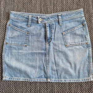 Blå jeanskjol  - Snygg ljusblå jeanskjol från Lindex med klassisk femficksdesign, bälteshällor och knappstängning framtill. Midjemått - ca 44 cm, Längd - ca 40 cm