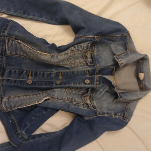 Klassisk blå jeansjacka, storlek 13/14 - Snygg blå jeansjacka från C&A Clockhouse i klassisk modell med knappar framtill och två bröstfickor. Jackan har kontrastsömmar och slitna detaljer som ger en cool vibe. Perfekt att slänga över en hoodie eller t-shirt.