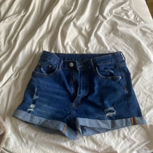 Mörkblå jeansshorts - Snygga mörkblå jeansshorts med uppvikta benslut och slitna detaljer framtill och baktill. Klassisk femficksmodell med dragkedja och knapp. Perfekta för sommaren och ger en avslappnad vibe. Pris kan diskuteras.