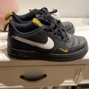 Nike Air Force 1 svart/gul 37.5 - Svarta Nike Air Force 1 med vita och gula detaljer. Skorna har svart skinn, vit swoosh och små gula accenter, samt gulrutiga dragflikar. Klassisk rund tå, snörning och platt sula. Perfekta för dig som vill sticka ut med en unik färgkombo.