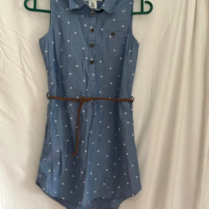 Blå ärmlös klänning H&M med bälte - Söt blå ärmlös klänning från H&M med vita blommiga prickar, skjortkrage och knappar framtill. Brunt flätat bälte i midjan och liten bröstficka. Tillverkad i 100% bomull, perfekt för varma dagar.