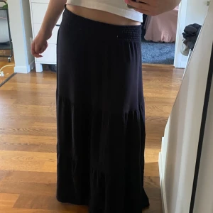 Svart långkjol med smock i midjan - En svart långkjol med smockad midja. Kjolen är luftig och har en lätt utställd form med diskreta volanger. Längden på kjolen är 92cm och längden på smocken är 7cm 