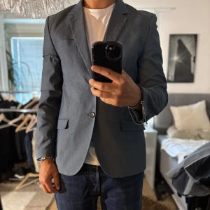 Blå kavaj från H&M, storlek 48 - Snygg blå kavaj från H&M i storlek 48. Klassisk modell med två knappar framtill, fickor med lock och fyra knappar vid ärmslutet. Perfekt för dig som vill ha en stilren och enkel look. Materialet är lätt och känns bekvämt på.