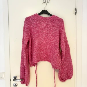 Stickad rosa tröja med öppen rygg - Trendig stickad rosa tröja med ballongärmar och öppen rygg med snörning. Snygg melerad look och croppad passform. Perfekt för dig som vill sticka ut med en cool detalj på ryggen och älskar färg.