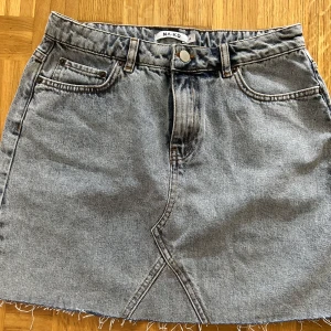 Grå jeanskjol från NA-KD, stl 40 - Snygg grå jeanskjol från NA-KD i klassisk femficksmodell. Kjolen har rå, fransig fåll och coola sömdetaljer framtill. Perfekt för dig som gillar en avslappnad och trendig stil. Passar till allt och är superenkel att styla.