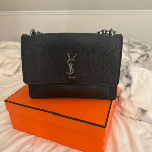 Svart YSL handväska i skinn - Säljer en svart handväska från YSL med silverfärgad logga och kedjerem. Väskan är i grynigt skinn och har flera fack, inklusive ett med dragkedja. Perfekt storlek för det viktigaste och riktigt snygg till alla outfits.