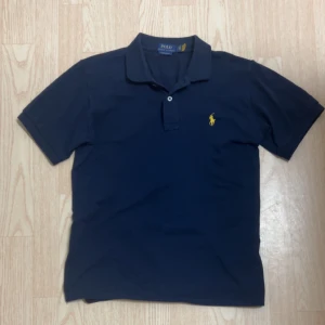 Ny Marinblå pikétröja Ralph Lauren S - Snygg marinblå pikétröja från Ralph Lauren i slim fit-modell. Klassisk krage, korta ärmar och gul broderad logga på bröstet. Tillverkad i bomull för en skön och luftig känsla. Perfekt för dig som gillar stilren och sportig look.
