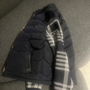 Mörkblå dunväst från Burberry M - Säljer en mörkblå dunväst från Burberry med klassiskt rutigt foder i beige, svart, vitt och rött. Västen har dragkedja framtill, två fickor med dragkedja och diskret Burberry-logga på ryggen. Perfekt för lager-på-lager och snygg detalj med rutigt mönster på insidan.