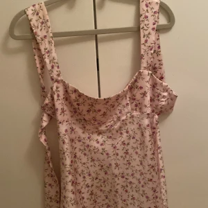 Ljusrosa blommig kortklänning med rosset i ryggen  - Supersöt ljusrosa kortklänning med blommigt mönster och breda axelband. Klänningen har en lätt utsvängd passform och är perfekt för varma dagar. Materialet känns mjukt och svalt, troligtvis polyester eller bomull. Perfekt för en somrig look!MAN SKA KNYTA ROSSET BAK