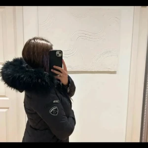 Svart lång pufferjacka med pälsluva - Säljer en svart lång pufferjacka från rock and blue med stor fluffig fuskpäls runt luvan. Jackan har quiltad design, dragkedja och knappar framtill samt en patch på ärmen. Perfekt för kalla dagar och riktigt snygg siluett. Liten skada (se bild)