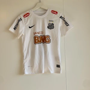 Santos Neymar Jr matchtröja Nike - Vit Santos FC fotbollströja från Nike med Neymar Jr och nummer 11 på ryggen. Tröjan har korta ärmar, svarta tryck och guldiga detaljer, samt flera sponsorer. Materialet är lätt och andas med Dri-FIT-teknologi. Matchande shorts med klubbmärke och nummer 11 ingår.