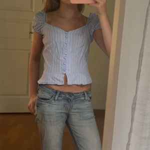 Blåvit randig blus från Dilvin Basic - Supersöt blus i ljusblått och vitt från Dilvin Basic. Den har puffiga korta ärmar, knappar framtill och en liten slits nertill. Snyggt figursydd med v-ringning och tunnt, luftigt material. I storlek 36 men passar xs-s