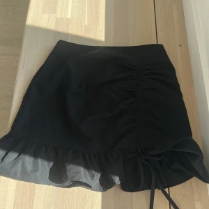 Svart kortkjol med volang och snörning - Supersöt svart kortkjol med volangkant och snörning framtill. Kjolen har en lätt rynkad effekt längs ena sidan och är figurnära med en feminin touch. Perfekt att styla med en crop top eller oversized hoodie för en trendig look.