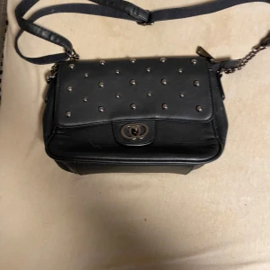 Svart axelväska med nitar - Snygg svart axelväska i skinnimitation med silverfärgade nitar på locket och kedjedetaljer på axelremmen. Väskan har ett vridlås framtill och en stilren, fyrkantig form. Perfekt för dig som gillar en edgy look.