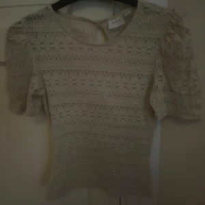 Beige spetsblus från Vila XS - Supersöt beige blus från Vila i storlek XS. Blusen har korta puffärmar och är helt täckt av vacker spets med blommigt mönster. Rund halsringning och knapp i nacken för en extra detalj. Perfekt för dig som gillar romantisk och feminin stil.