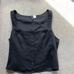 Svart linnekavaj med knappar - Stilren svart linnetopp med breda axelband och snygg knappdetalj framtill. Toppen har en figurnära passform och är perfekt att matcha med jeans eller kjol för en trendig look.