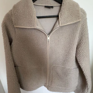 Beige teddyjacka från 157, XS - Mysig beige teddyjacka från 157 i storlek XS. Jackan har dragkedja framtill, två stora fickor och en hög krage. Perfekt för kyliga dagar när du vill vara både varm och snygg. Fleece-liknande material som känns supermjukt.