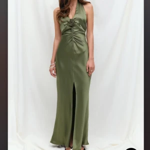 Olivgrön långklänning i satin - Snygg olivgrön långklänning i glansig satin med halterneck och öppen rygg. Klänningen har en dekorativ blomdetalj framtill, smickrande rynk vid midjan och en slits framtill för extra edge. Perfekt för dig som vill sticka ut med en elegant och modern look.