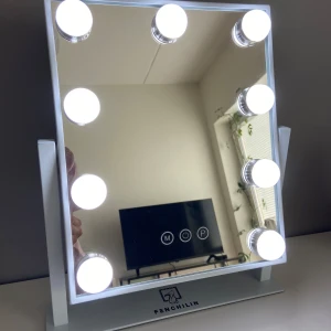 Fenchilin Hollywood sminkspegel -  Sminkspegel från Fenchilin med vit ram och 9 stora LED-lampor runt spegeln. Touchknappar på spegeln för att justera ljusstyrka och färgtemperatur. D ett finns tre ljus temperaturer: vitt, gult och blått. Spegeln står stadigt på en metallbas och kopplas in med sladd på baksidan. Höjd: 30 cm Bredd: 25 cm Djup: 9 cm