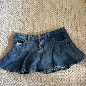 Blå jeanskjol med volangkant - Säljer en blå jeanskjol i kort modell med volangkant nertill. Kjolen har klassiska jeansdetaljer som fickor fram och bak, bälteshällor och knappstängning. Perfekt för dig som gillar en avslappnad och trendig stil. Den har även innershorts. 