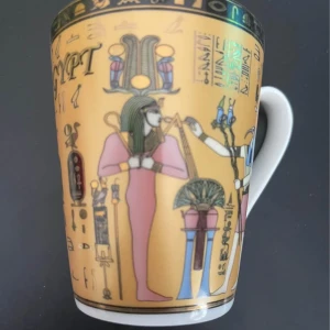 mugg - Bilderna visar en mugg med motiv från forntida Egypten, inklusive hieroglyfer och färgstarka illustrationer av egyptiska gudar och faraoner. Muggen har detaljerade och dekorativa mönster som ger en känsla av historia och mystik. Ingen bok syns på bilderna.