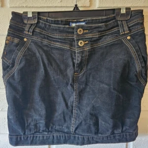 Mörk jeanskjol från Blue Forest - Snygg svart/grå jeanskjol från Blue Forest med coola kontrastsömmar, dubbla knappar fram och klassiska fickor både fram och bak. Kjolen har bälteshällor och en kort, rak passform som ger en avslappnad men trendig look.  Ett litet trådsläpp på sidan, men inget hål.  Har legat nerpackad något år