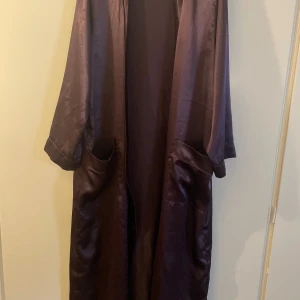 Ella May, Lindex lila/plommon morgonrock, storlek 38/40  - Snygg morgonrock i mörklila från Ella May för Lindex. Morgonrock har en loose passform, vida ärmar och stora fickor framtill. Tillverkad i glansigt satinliknande material som ger en lyxig känsla. Perfekt för dig som gillar stilrena och bekväma plagg.