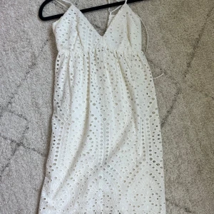 Vit broderad sommarklänning med spets - Superfin vit klänning med broderade detaljer och spetsmönster. Klänningen har smala axelband, v-ringning och omlottkänsla upptill. Den är kort och har en vågig kant nertill. Perfekt för varma dagar och ger en romantisk vibe. Storlek Xs men går att justera så att den massa alla storlekar. Märke H&M. 