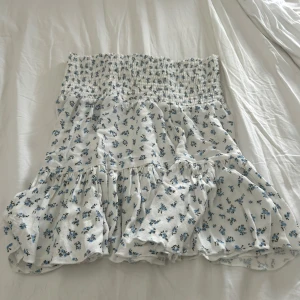 Vit blommig kjol.  - Supersöt vit kortkjol med små blå blommor och bred smock i midjan. Kjolen har volangiga lager som ger en flowy känsla och passar perfekt till varma dagar. Lätt och luftig, perfekt för en chill sommarlook. Storlek S.