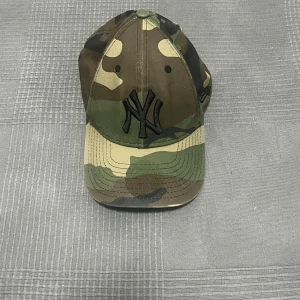NY Yankees keps i camouflage från New Era - Snygg New York Yankees keps från New Era i klassiskt 9FORTY-snitt. Kepsen har ett grymt camouflage-mönster i grönt, brunt och beige samt svart broderad NY-logga framtill. Justerbar rem baktill för perfekt passform. Perfekt för dig som vill sticka ut med streetstyle.                       Inre omkrets max 50cm.                                                        Frågor? Hör av dig