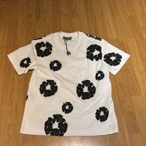 Denim tears T-Shirt  - Vit t-shirt med unikt svart mönster över hela plagget. Klassisk passform med rund hals och korta ärmar. Tillverkad i mjuk bomull som känns skön mot huden. Perfekt för dig som gillar statement-plagg och coola prints.