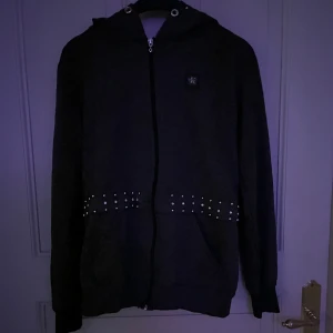 Svart hoodiejacka Calvin Klein  - Svart hoodiejacka från Calvin Klein Jeans med dragkedja och coola silvriga nitar runt midjan och på huvan. Jackan har en enkel, clean look med långa ärmar och fickor framtill. Pris kan diskuteras. 