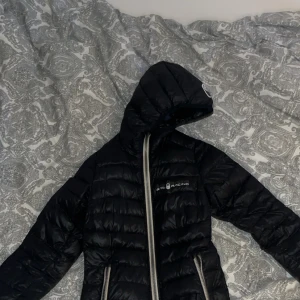 Svart pufferjacka från Sail Racing - Svart pufferjacka från Sail Racing med huva och tvåvägsdragkedja i silver. Jackan har quiltad design och logga på bröstet. Perfekt för kalla dagar och har en riktigt clean look. Snygg passform och funkar till många outfits.