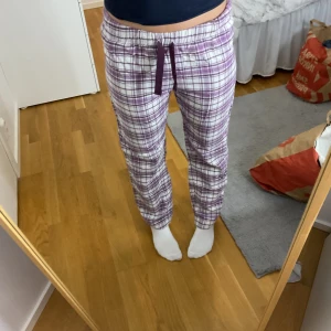 Lila-rutiga mjukisbyxor med snörning - Mysiga mjukisbyxor i vitt och lila rutigt mönster. Byxorna har en relaxed passform, elastisk midja och lila snörning framtill. Perfekta för chill dagar hemma. Tillverkade i mjukt bomullstyg för extra komfort.