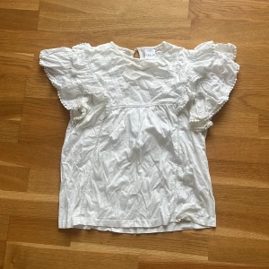 Vit blus med volangärmar från Zara - Supersöt vit blus från Zara med volangiga korta ärmar och broderade detaljer. Blusen har rund halsringning, knapp i nacken och små bollar längs ärmsluten. Perfekt för en fräsch och romantisk look!