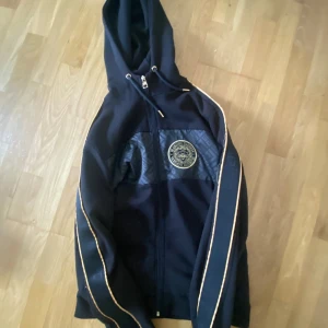 Svart zip hoodie  från Glorious Gangsta - Svart zip hoodie med huva från Glorious Gangsta. Jackan har guldfärgade detaljer längs ärmarna, dragkedja framtill och ett broderat emblem på bröstet. Snyggt mönstrad panel över bröstet och snörning i huvan. Perfekt för dig som vill sticka ut med stil.