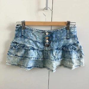 Blå volangkjol i jeans från Terranova - Supersöt kort jeanskjol från Terranova med volanger och snygga knappar framtill. Kjolen har ljusblå tvättad denimlook och bälteshällor i midjan. Perfekt för dig som vill ha en cool och avslappnad stil med extra edge. En av knapparna har gått sönder