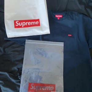 Mörkblå Supreme Box Logo t-shirt - Supreme t-shirt i mörkblått med klassisk röd box logo broderad på bröstet. T-shirten är i bomull och har en normal passform med korta ärmar och rund hals. Perfekt för dig som gillar streetwear och vill ha en clean look med ikonisk Supreme-vibe.