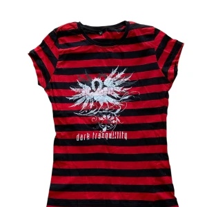  Dark Tranquillity randig t-shirt - Röd och svart randig t-shirt från Continental med tryck av Dark Tranquillity och en stor vit grafisk design framtill. T-shirten har korta ärmar, rund hals och är gjord i mjuk bomull. 