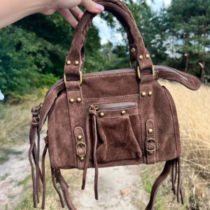 Brun mocka city väska med fransar - Snygg brun handväska i mocka med coola fransdetaljer och guldfärgade nitar. Väskan har både handtag och axelrem, dragkedja upptill samt en liten ficka med dragkedja framtill. 