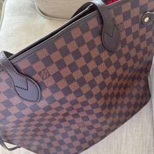 Axelväska brun - Säljer en axelväska i brunt Damier-mönster. Väskan har långa handtag, detaljer i mörkbrunt skinn och guldiga metalldetaljer. Insidan är röd med svarta ränder och har en innerficka med dragkedja. Perfekt för dig som gillar lyxig och tidlös design.