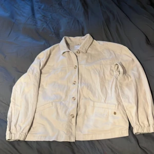 Beige overshirt från Pernilla Wahlgren - Snygg beige overshirt från Pernilla Wahlgren Design. Jackan har klassisk krage, knäppning framtill, två fickor och elastiska muddar vid ärmslut och nederkant. Tillverkad i bomull för en skön och avslappnad vibe. Perfekt att slänga över en hoodie eller t-shirt.