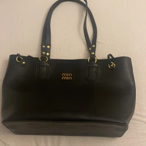 Svart handväska från Miu Miu i skinn - Snygg svart handväska från Miu Miu i skinn med dubbla axelband och guldfärgade metalldetaljer. Klassisk och rymlig modell med stilren design och logga framtill. Perfekt för dig som vill ha en elegant och tidlös väska med lyxig känsla.
