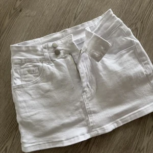Vit jeanskjol med fickor och dragkedja - Snygg vit kort jeanskjol med klassiska fem fickor, dragkedja och dubbla knappar framtill. Perfekt för en fräsch och stilren look. Kjolen har bälteshällor och raka linjer som ger en clean vibe.