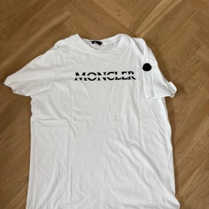 Vit Moncler t-shirt med logga - Vit t-shirt från Moncler i 100% bomull med svart Moncler-tryck på bröstet och svart patch på ärmen. Klassisk passform och rund halsringning, perfekt för en clean och stilren look.