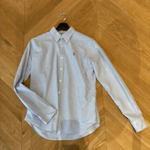 Ljusblå skjorta från Ralph Lauren - Snygg ljusblå skjorta från Ralph Lauren i slim fit-modell. Skjortan har klassisk button down-krage, långa ärmar och knäppning framtill. Sitter som en XS. Broderad logga på bröstet ger en clean och stilren look. Perfekt för dig som gillar preppy stil. Pris kan diskuteras