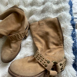 Beiga mockaboots med nitar från Zadig & Voltaire - Snygga boots i beige mocka från Zadig & Voltaire med coola mässingsfärgade nitar och ringdetalj runt ankeln. De har rund tå, platt sula och dragkedja på sidan. Perfekta för dig som gillar en avslappnad men edgy stil. I storlek 38. Mina absoluta favoritskor som passar till ALLA tillfällen, tyvärr har de blivit för små för mig🥹