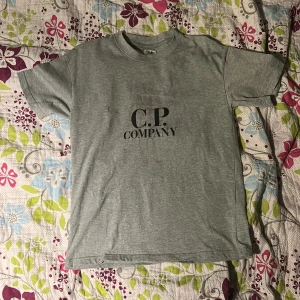 Grå t-shirt från C.P. Company - Snygg grå t-shirt från C.P. Company med svart logga tryckt på bröstet. Klassisk rund halsringning och korta ärmar. Tillverkad i mjuk bomull som känns skön mot huden. Perfekt för en clean och stilren look.